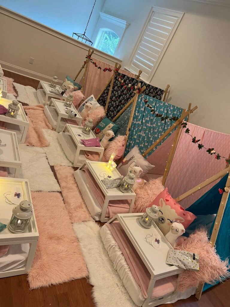 A row of teepee tents set up indoors with pillows, blankets, and plush toys. Each tent has a small table in front with decor items and a lantern. Perfect for a slumber party or as a unique party rental option, the room features a hardwood floor and rugs.