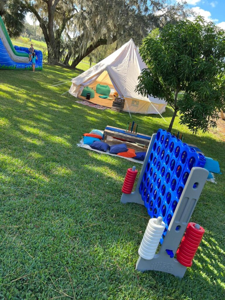 A large outdoor game and pillows are set on grass near a white tent with bean bags inside, perfect for a teepee-themed slumber party. In the background, a colorful inflatable slide adds fun to the scene, while trees and sunlight create a serene atmosphere.