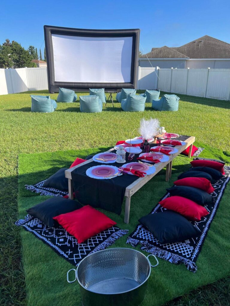 Outdoor movie setup in a backyard with an inflatable screen, bean bag seating, and a low table with red and black pillows for dining. Perfect for a party rental event, transforming your space into a whimsical getaway.