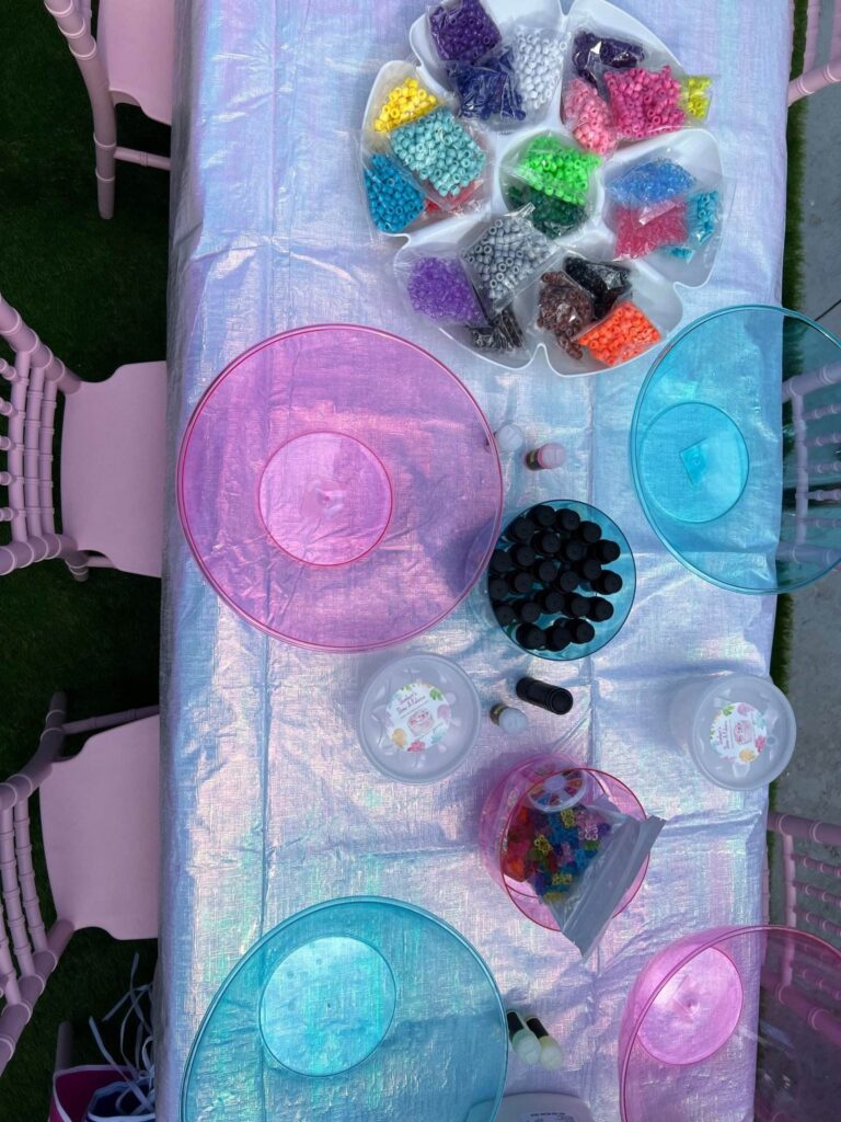 A table set up for a craft activity with various colored beads, clear containers, and colorful bowls on a shimmering tablecloth, perfect for a kids' party rental.