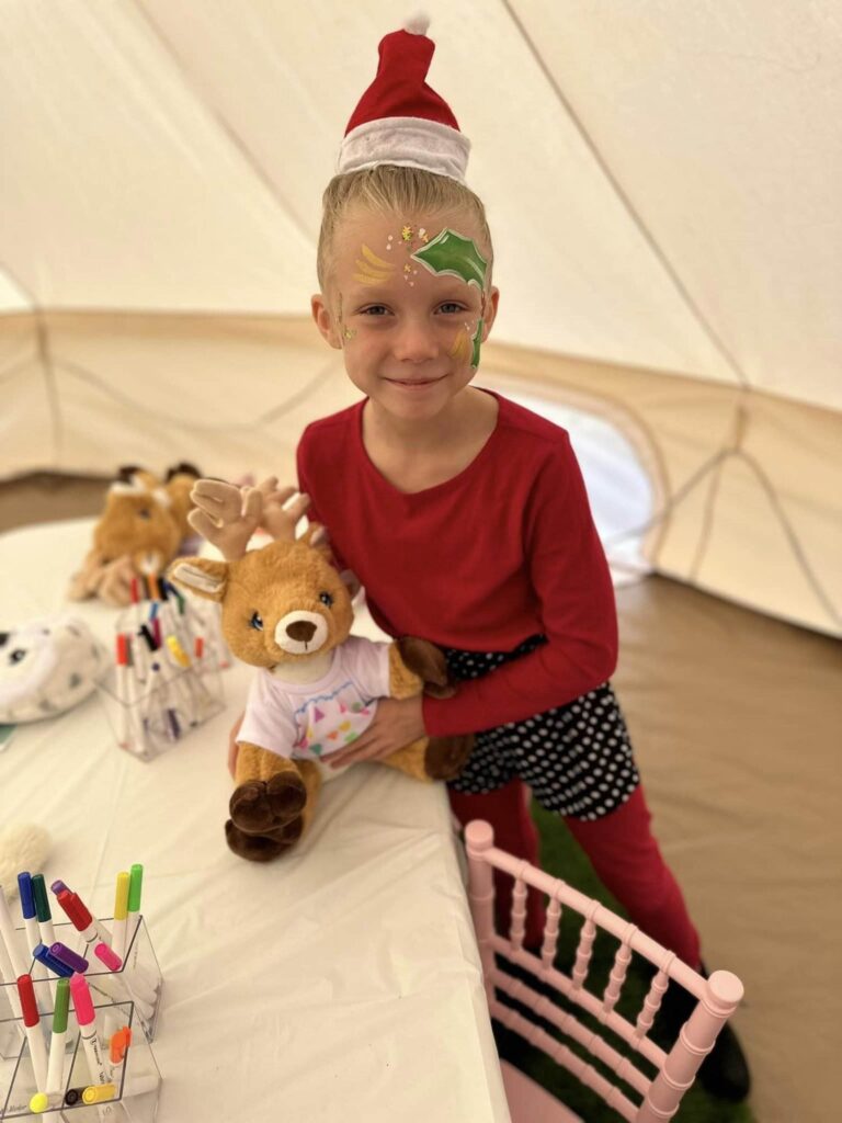 A child with face paint and a festive hat smiles while standing in front of a table with various craft supplies, stuffed animals, and exciting slime party materials.