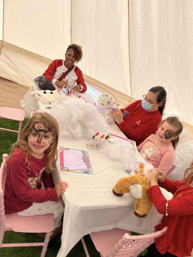 A group of children and adults sit around a table in a tent, engaging in a stuffed animal workshop. One child has face paint, and another wears a mask. Arts and crafts supplies are scattered on the table, creating an atmosphere reminiscent of a lively slime party.
