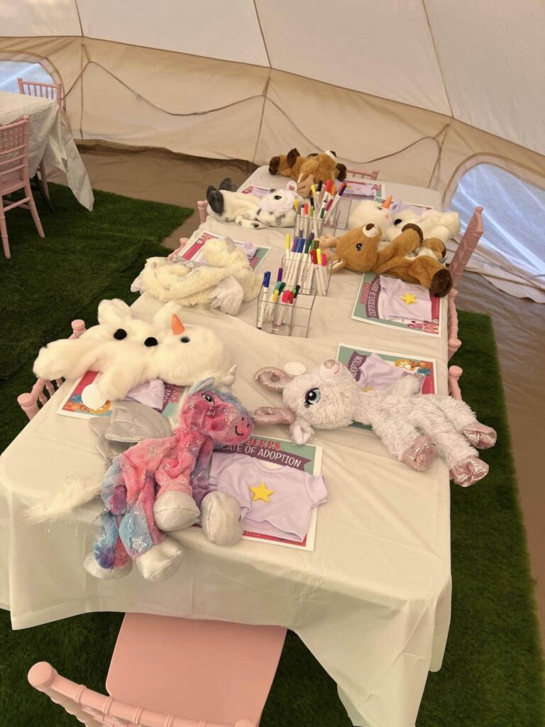 A long table with stuffed animals, coloring supplies, and adoption paper forms is set up in a white canopy tent. Pink chairs are arranged around the table, adding a charming touch to this kids party rental setup.