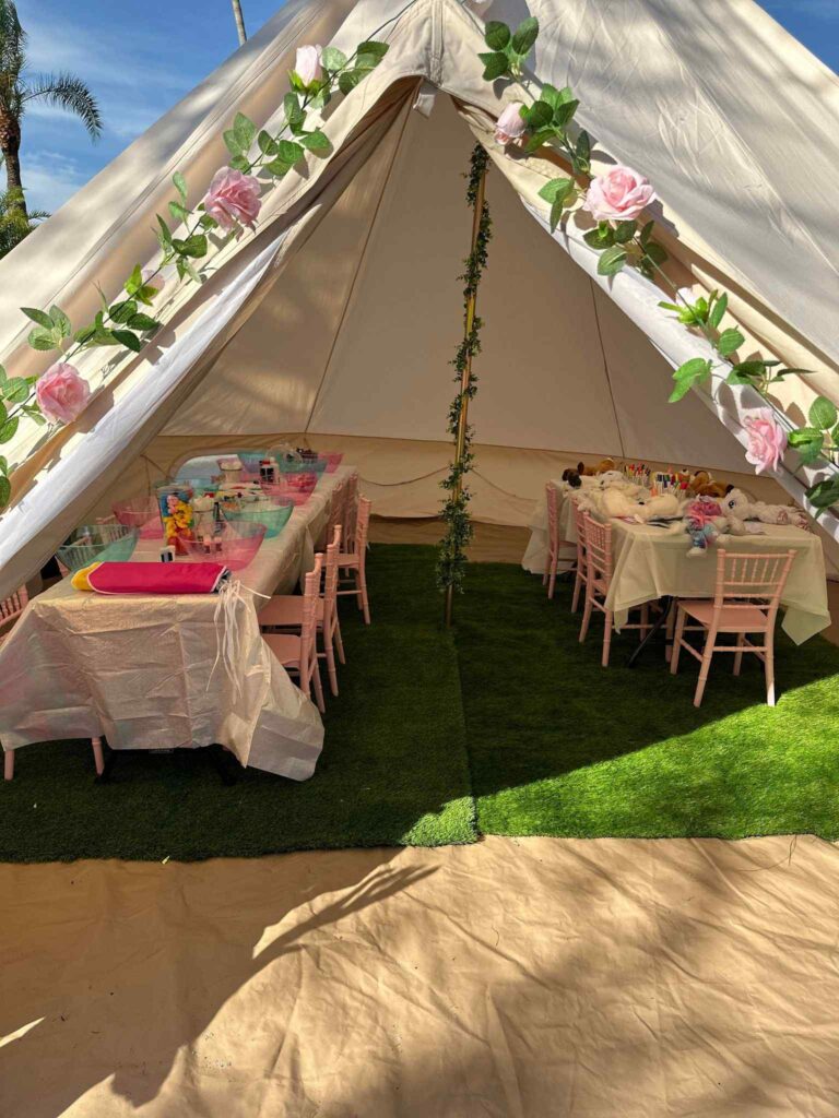 A decorated tent with tables and chairs set up for a kids party rental features pink plates, colorful cups, and flower garlands inside.