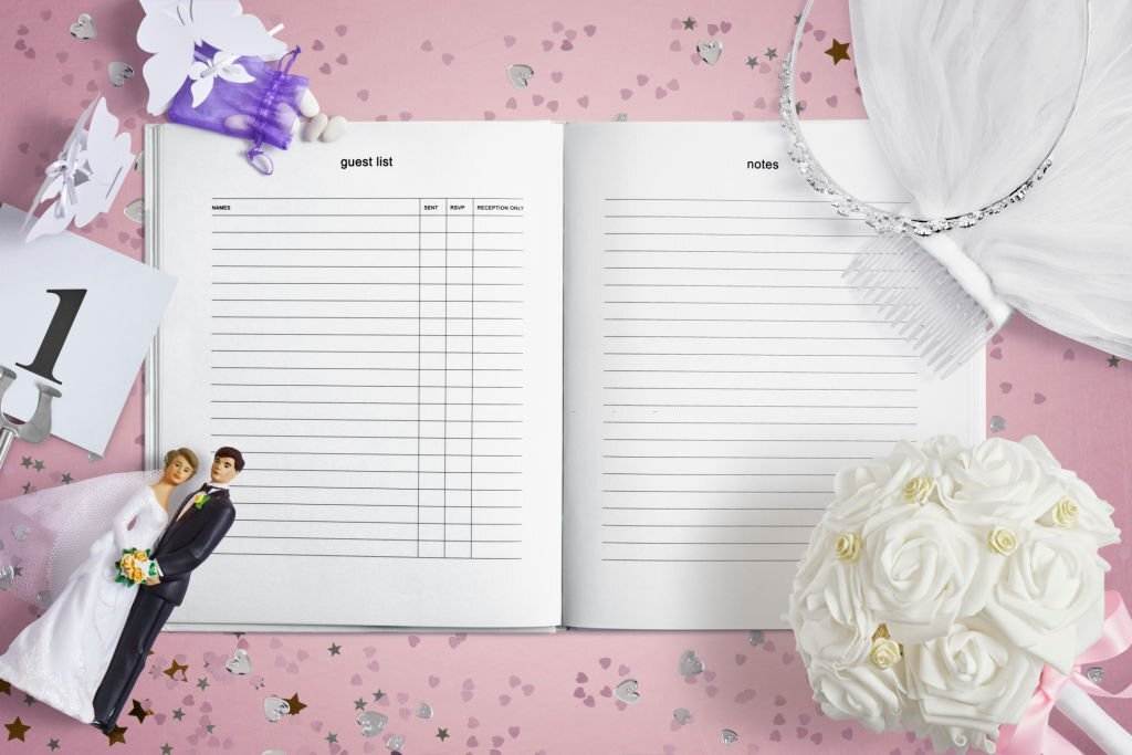 Open wedding planner on a pink confetti-strewn table, featuring sections for guest list and notes, flanked by bride and groom figurine, bouquet of white roses, and wedding decor pieces from our party service.