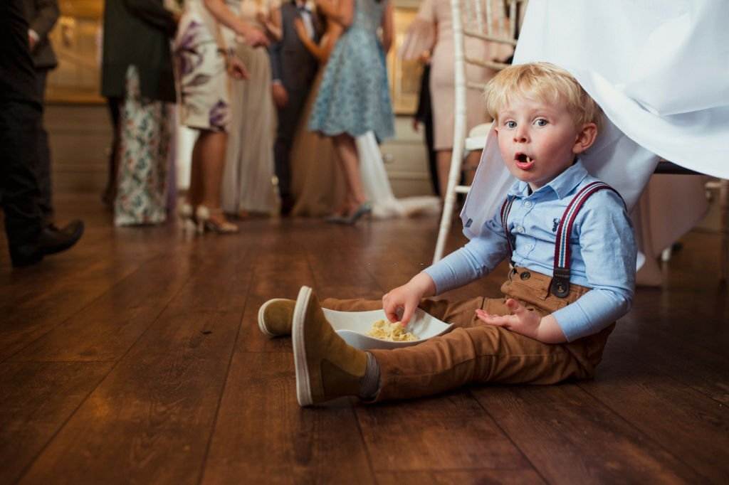 A child with blond hair is sitting on a wooden floor under a table, eating from a bowl, while adults chat in the background. The cozy setup reminds one of a slumber party with fun decorations scattered around.