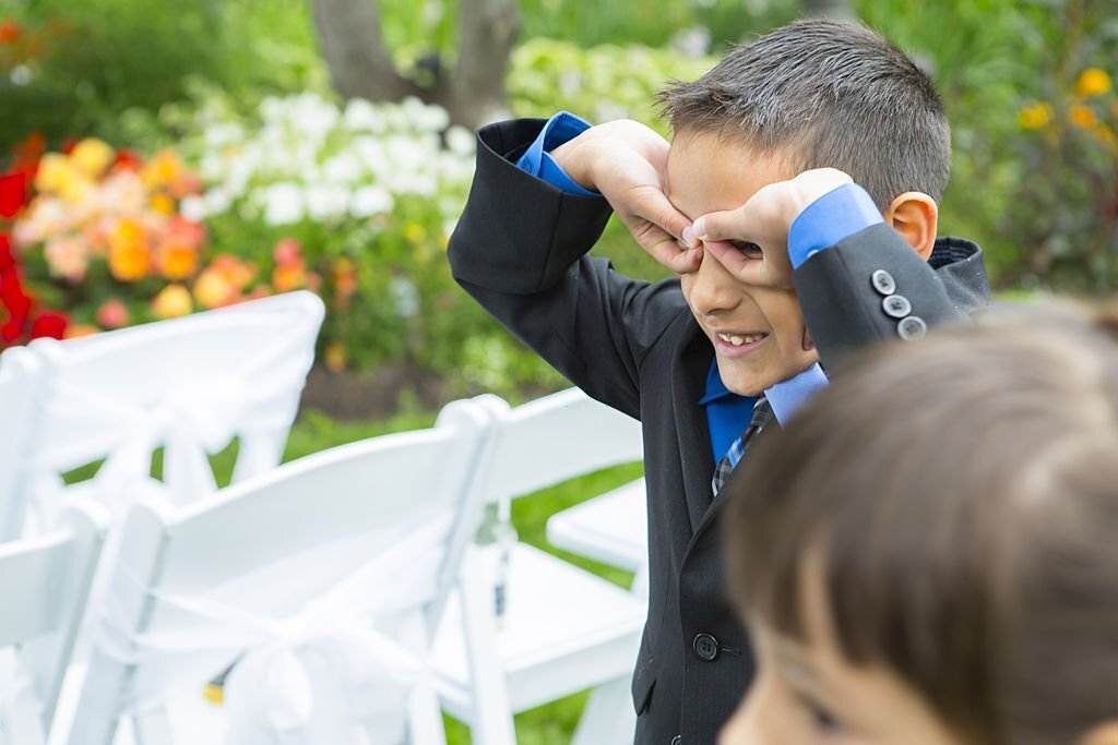 A young boy in a suit playfully makes binocular shapes with his hands over his eyes while standing in front of white folding chairs, colorful garden flowers, and a charming teepee from the party rental.