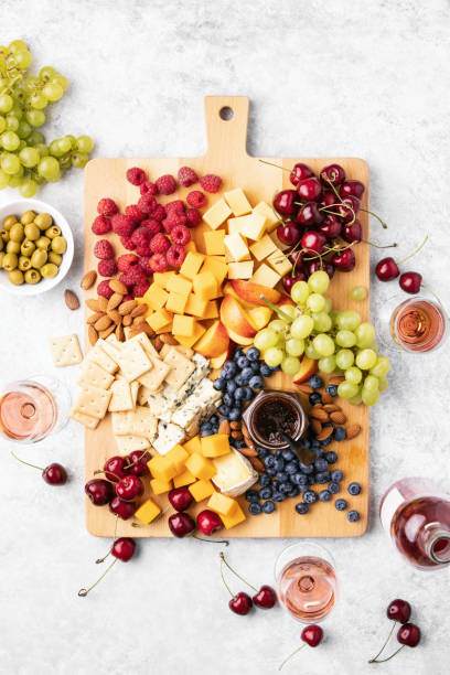 A wooden board with assorted cheeses, raspberries, grapes, cherries, blueberries, crackers, almonds, and olives sits on a light-colored surface. Glasses of rose wine surround the spread—a delightful setup perfect for any kids party rental or gathering.