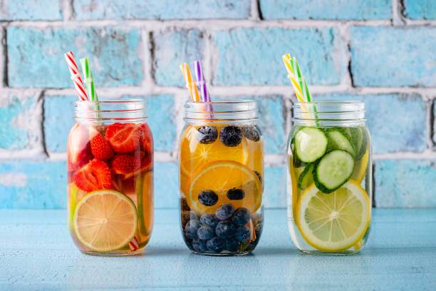 Three mason jars filled with colorful fruit-infused water: strawberries and lemon, blueberries and orange, cucumber and lemon. Each jar has patterned straws. Perfect addition to any party rental setup against a blue brick background.