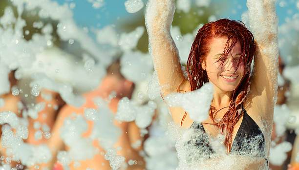Woman smiling in a black bikini top, enjoying a foam party outdoors, with party rental equipment and other people covered in foam in the background.