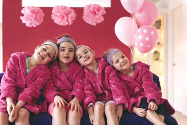 Four children in pink robes and matching headbands sit close together on a couch, smiling. Pink balloons and decorations are in the background, creating a cozy scene of a slumber party complete with fun teepee setups from the latest party rental service.