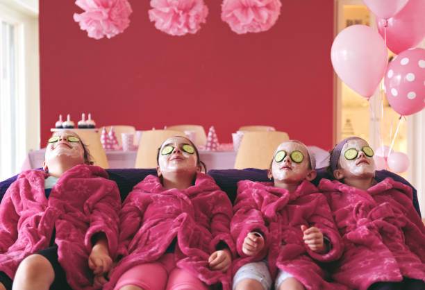 Four children wearing pink robes, facial masks, and cucumber slices on their eyes, lounging on a couch with balloons and party decorations in the background. This delightful scene showcases the perfect kids party rental setup for an unforgettable celebration.