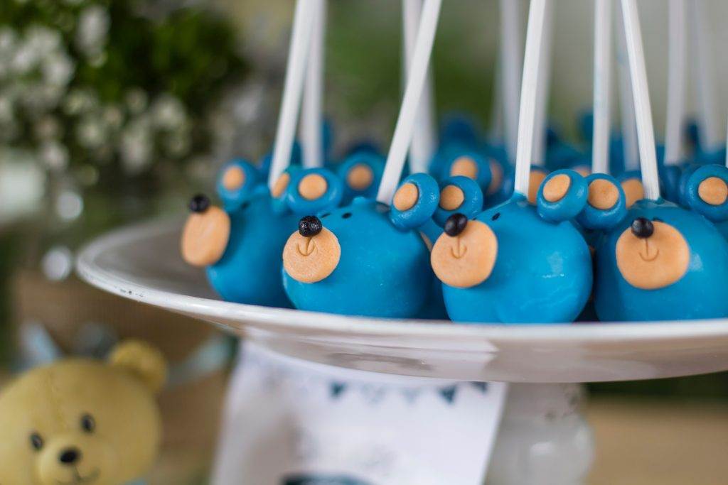A plate of blue bear-themed cake pops with small ears and orange snouts, displayed vertically on sticks. A blurred plush bear is visible in the background, perfect for a kids party rental setup.