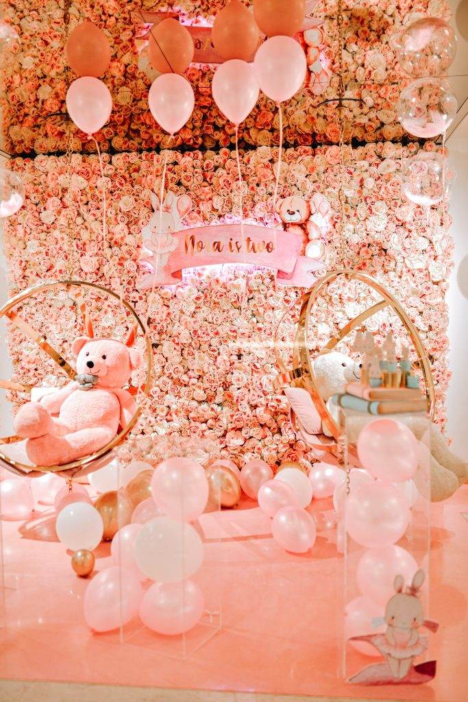 A pink and white floral wall with balloons, stuffed teddy bears in circular swings, and a banner that says "no act is too small." Pink and white balloons are scattered on the floor, perfect for a whimsical kids party rental.