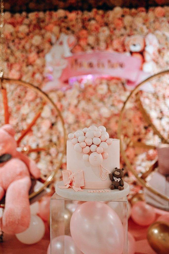 A pink birthday cake adorned with bubble decorations and a teddy bear figurine is placed on a glass stand. Perfect for a slime party, the backdrop features a floral wall, two large pink teddy bears, and balloons. Ideal for kids party rental or even a slumber party!