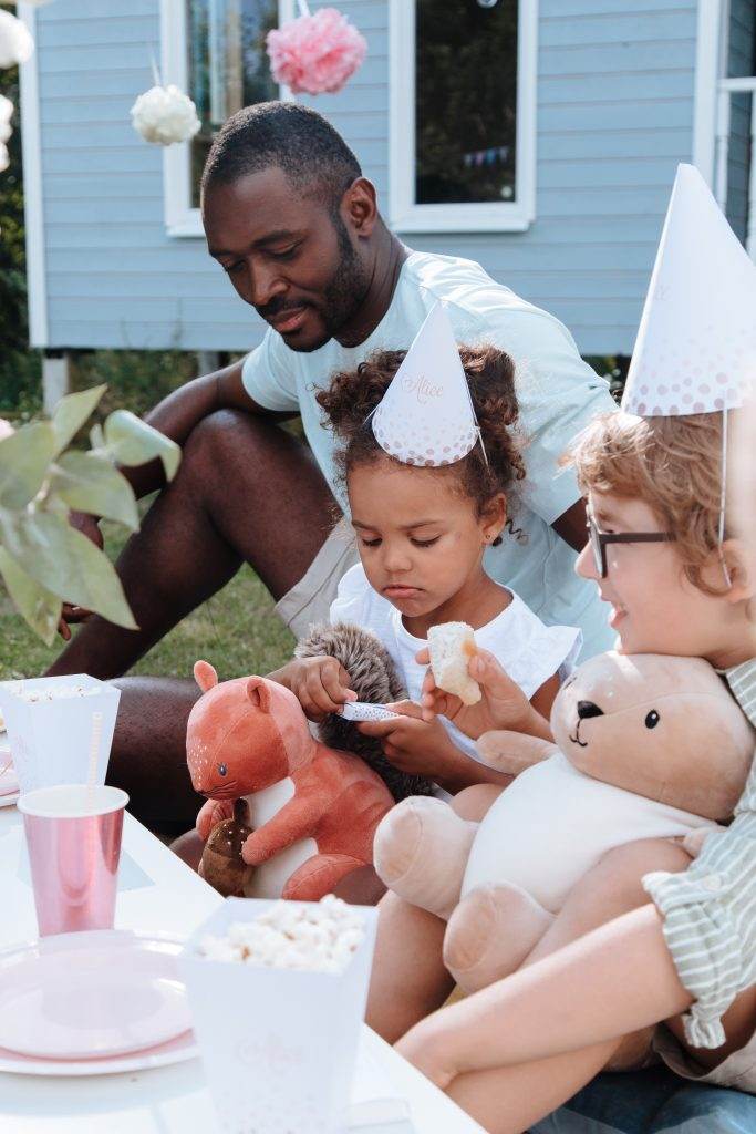A man and two children with party hats sit outdoors. One child is holding a teddy bear and the other is playing with a toy dinosaur. Cups and popcorn are on the table, creating a joyful scene that's perfect for any family gathering or party rental setup.