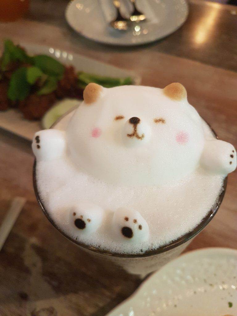 A latte features 3D latte art depicting a bear with eyes closed and arms extended, as if it's just waking up from a magical slumber party. In the background, a dish with food and utensils on a plate is visible.