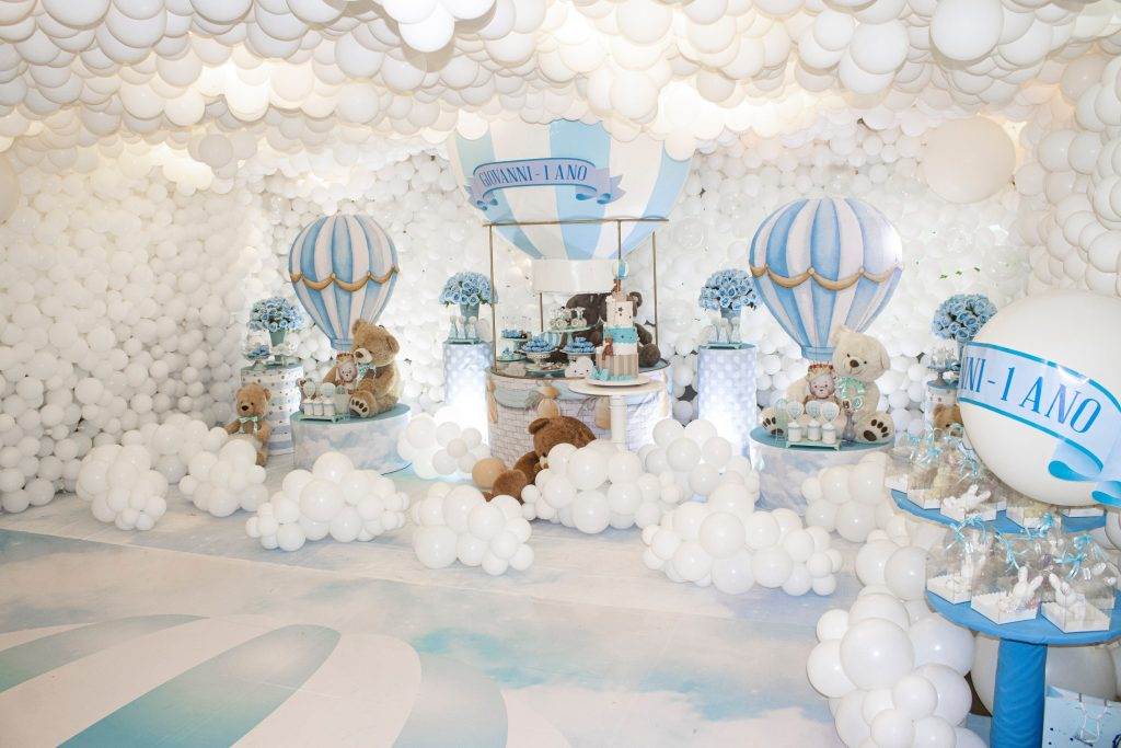 A room decorated with white balloons and blue accents features teddy bears, a teepee, a dessert table, and a sign that reads "Giovanni 1 Ano," indicating a charming first birthday celebration.