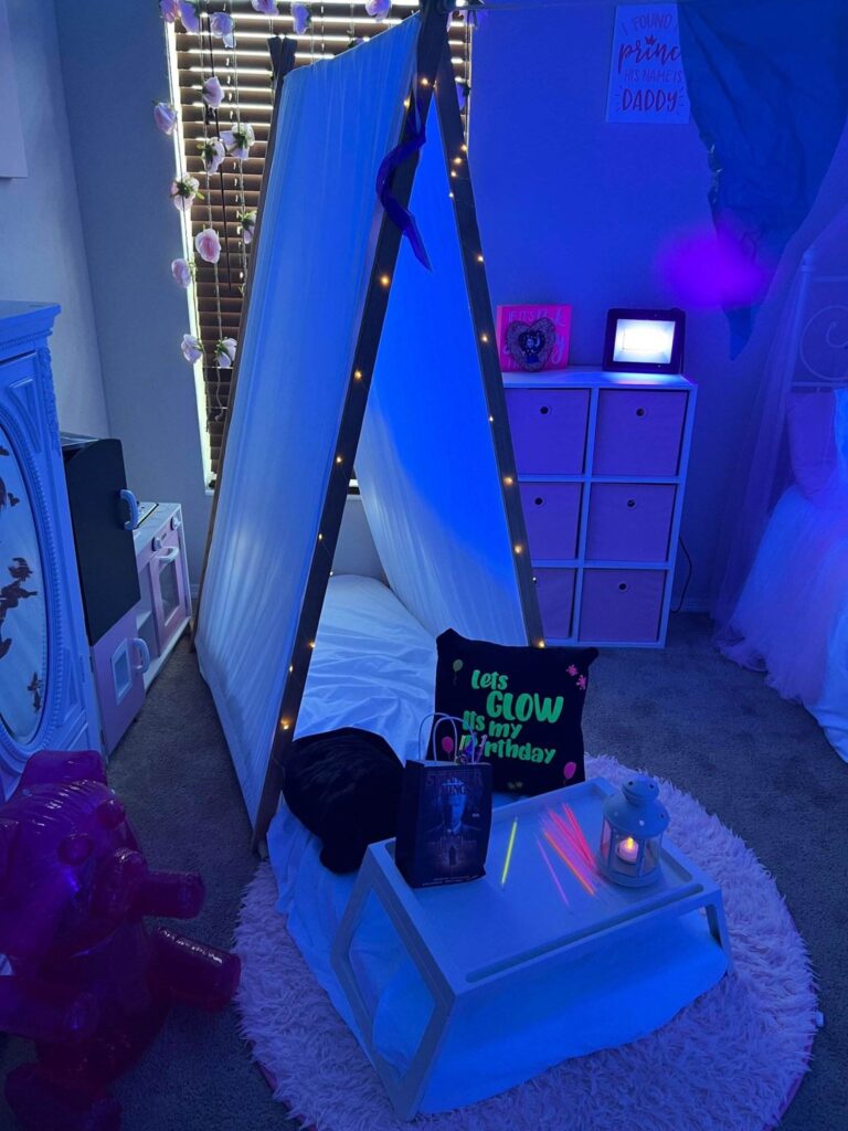 A child's bedroom with a teepee tent illuminated by string lights, surrounded by whimsical decorations and a slumber party-themed setup.