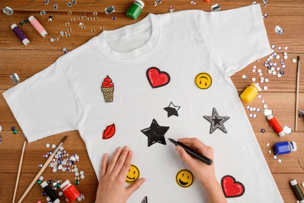 Hands decorating a white t-shirt with colorful fabric paint and appliques, including smiley faces and stars, at a slumber party on a craft-covered table.