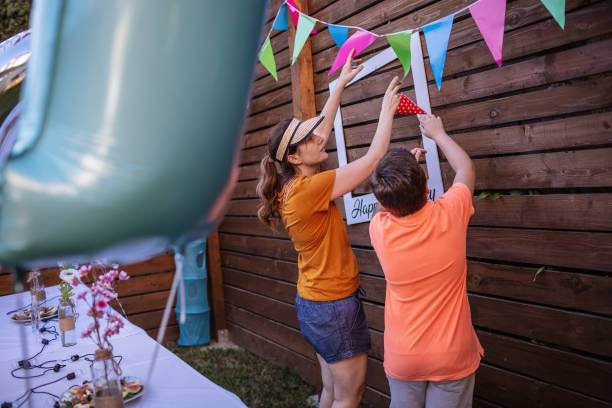 Two people hanging colorful bunting on a wooden fence at a slumber party, with a table of food and balloons visible.