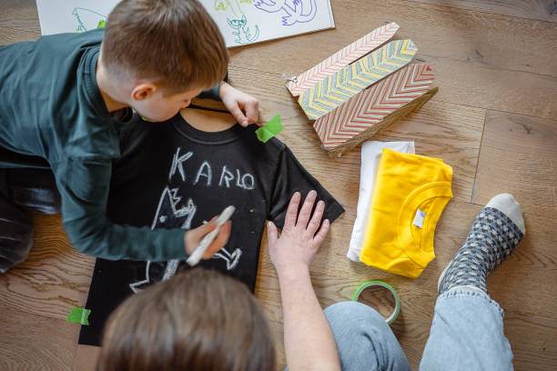 Adult helping a child decorate a black t-shirt with chalk for slumber party crafts, surrounded by gift boxes and a yellow shirt on a wooden floor.
