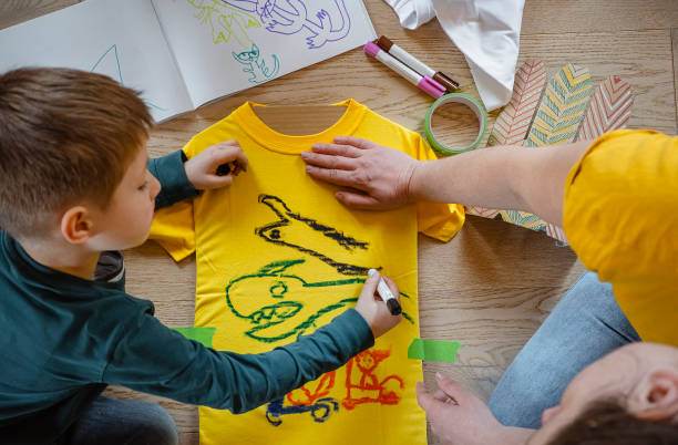 A child and an adult decorating a yellow t-shirt with fabric markers on a wooden table surrounded by art supplies for a slumber party craft.