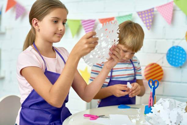 A girl and a boy crafting with paper and scissors at a slumber party, seated at a table decorated with colorful bunting.
