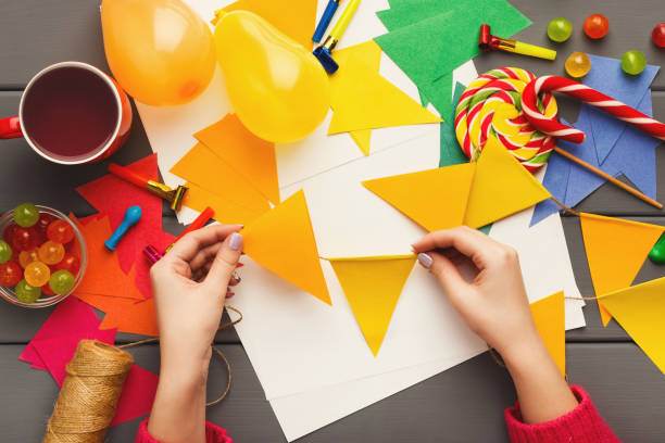 Hands engaged in slumber party crafts with colorful paper, scissors, and decorations on a table with candy, balloons, and a cup.