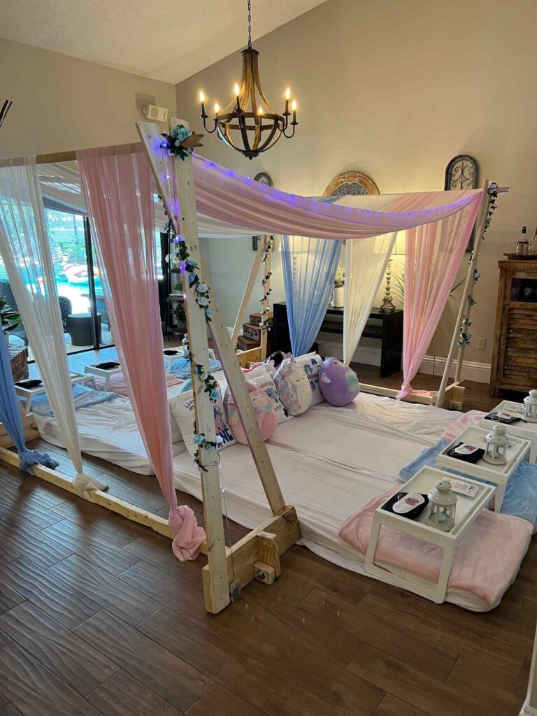 A cozy indoor canopy bed decorated with pink and blue curtains, fairy lights, and surrounded by decorative pillows and a small tray with a teapot, perfect for an ultimate canopy slumber.