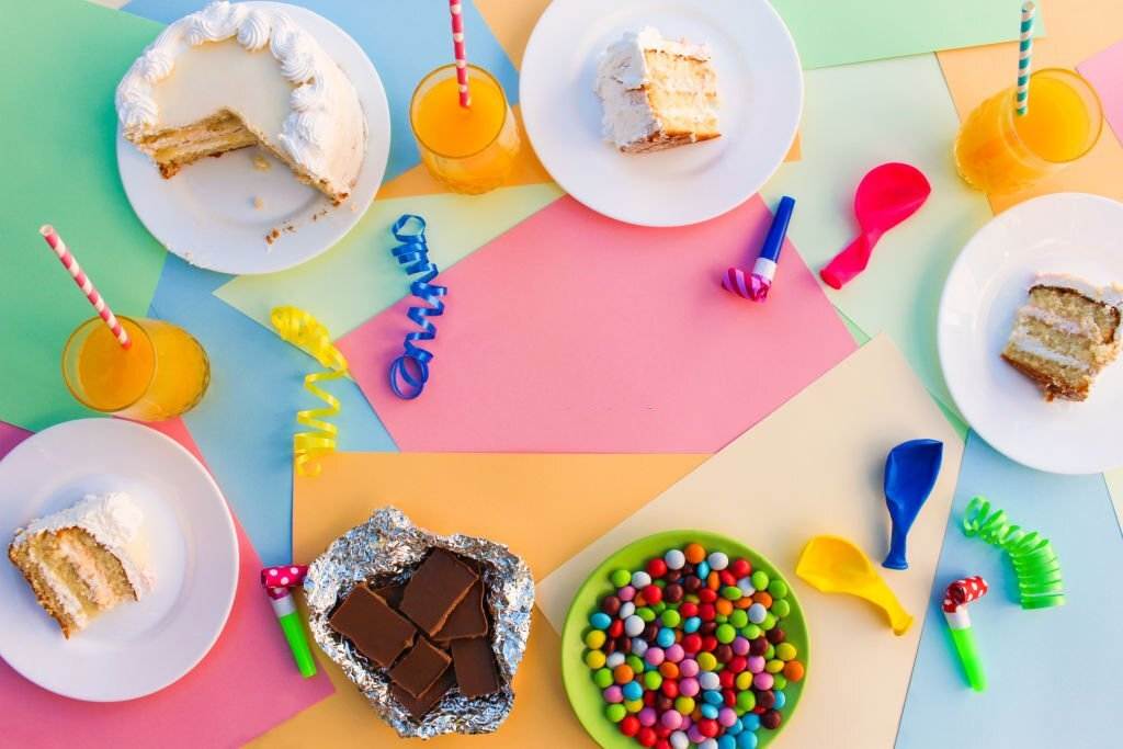 Top view of a colorful canopy slumber party table with sliced cake, drinks, and candies on a multicolored background.