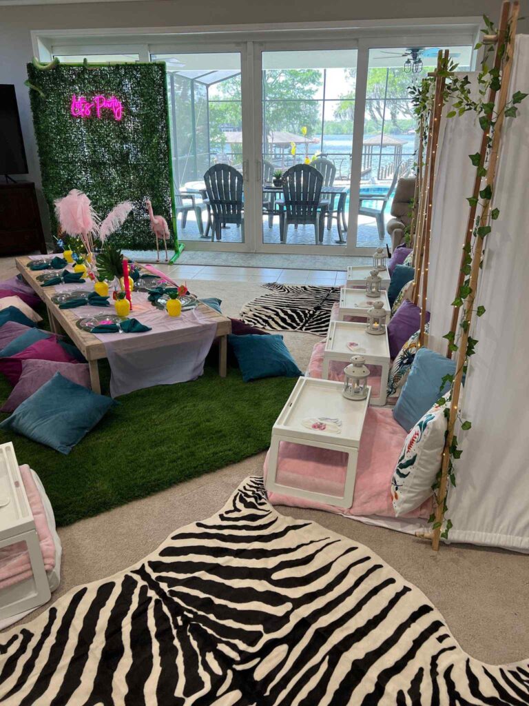 A vibrant indoor birthday party setup featuring a grass-like carpet, a long table with assorted colorful cushions, and a pool visible through glass doors.