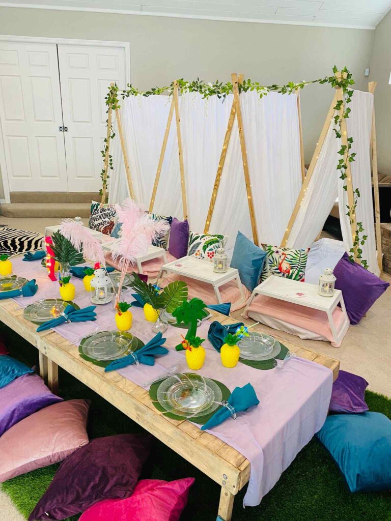 An indoor bohemian-style birthday party setup with a low wooden table, colorful cushions, and decorative items including pineapple centerpieces and hanging greenery.
