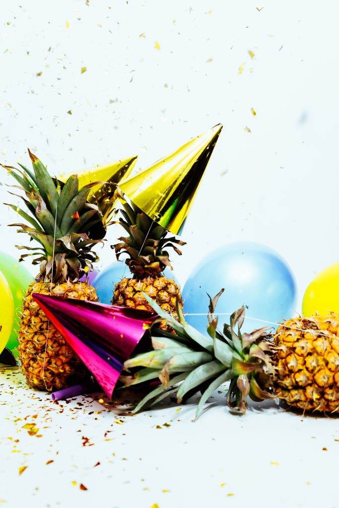 Two pineapples wearing party hats surrounded by colorful balloons and confetti on a white background, celebrating a tropical birthday party.