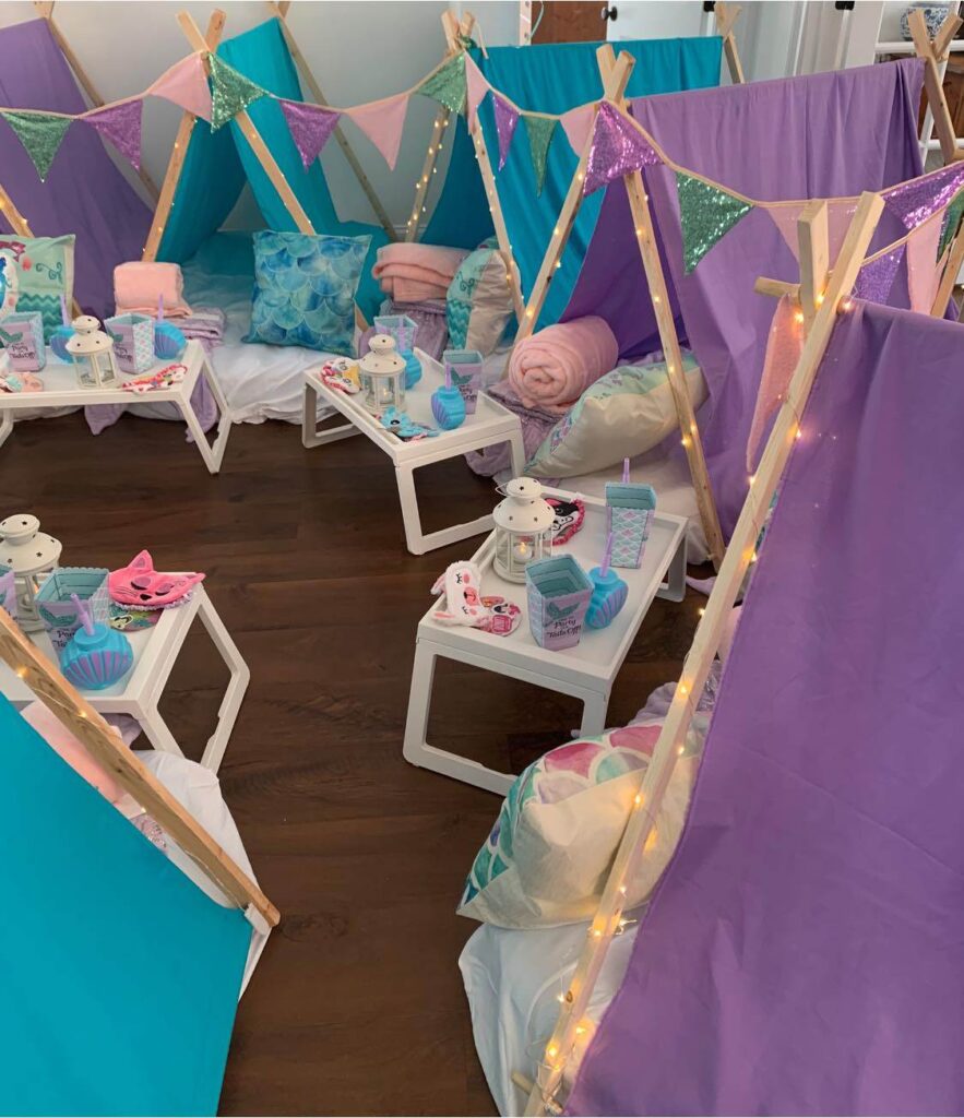 Indoor children's party setup with small teepees decorated with fairy lights, and tables set with teacups and saucers.