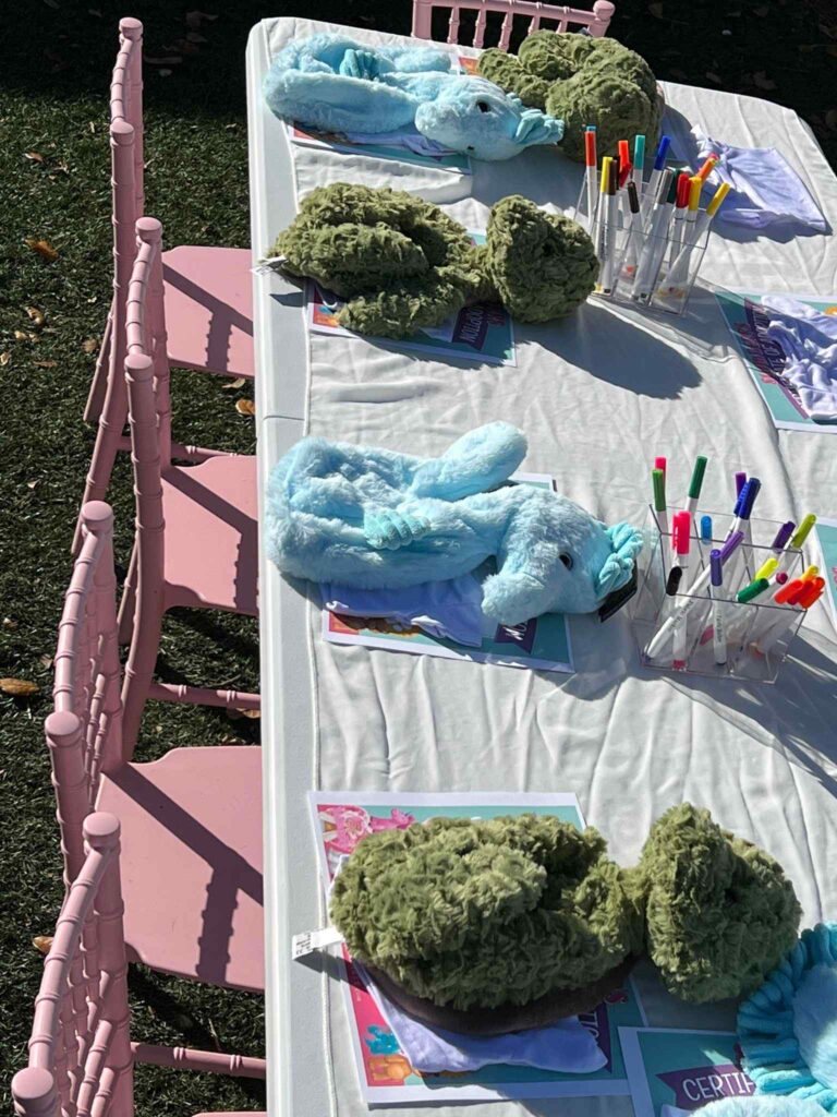 Outdoor table displaying plush crocodile toys and pens for coloring at a children’s event, featuring ultimate party upgrade with a pink fence in the background.