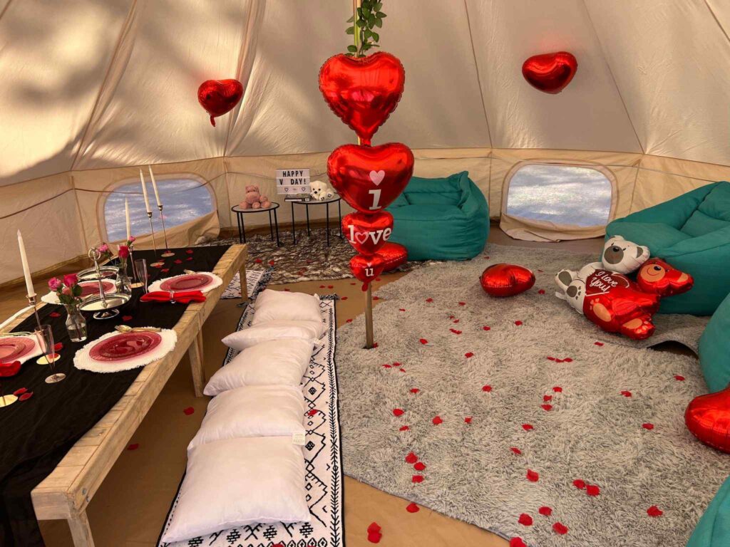 A unique slumber party setup with a dining area, red heart-shaped balloons, plush cushions, and rose petals scattered on the floor.