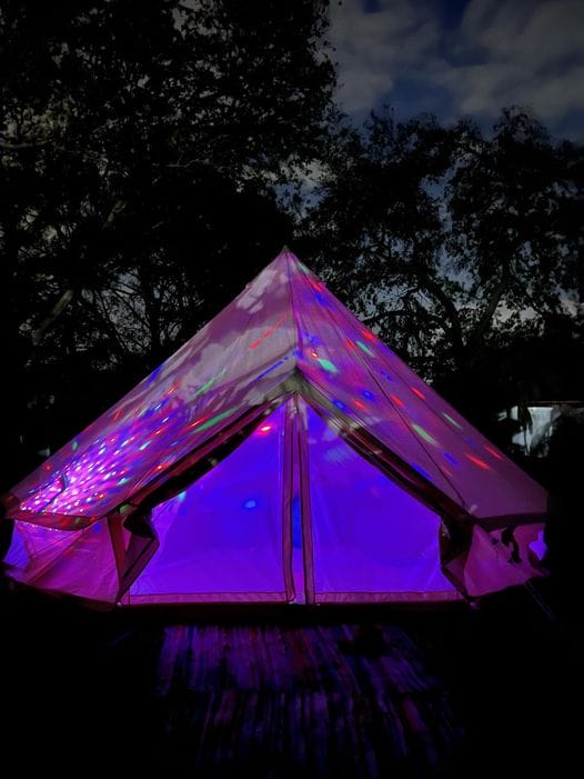 A tent illuminated by vibrant purple and pink lights under a twilight sky hosts a unique slumber party.