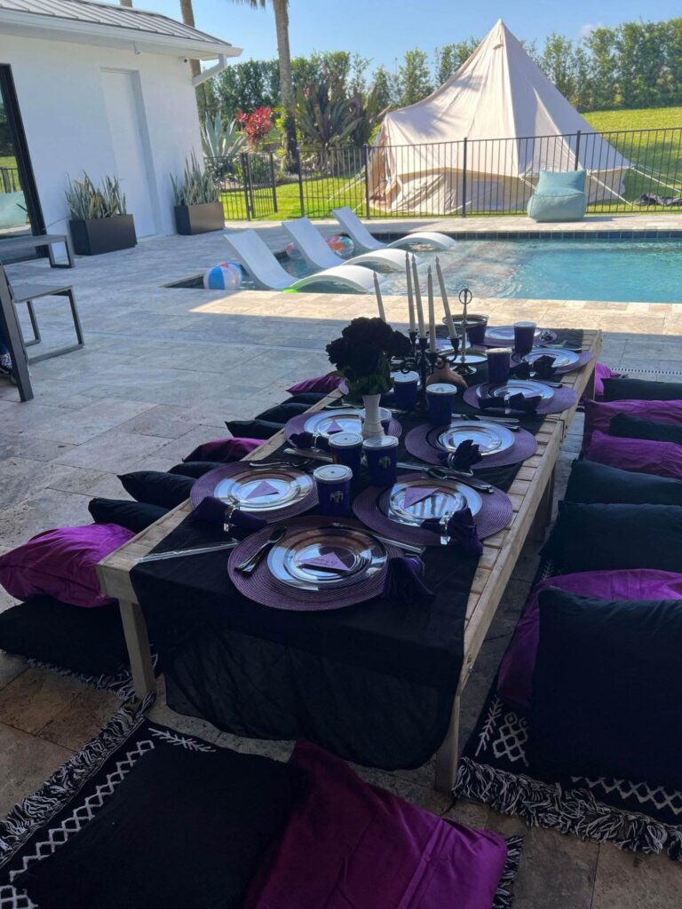 Outdoor dining setup for a birthday event by a pool with a long wooden table, black and purple cushions, complete tableware, and a tent in the background.