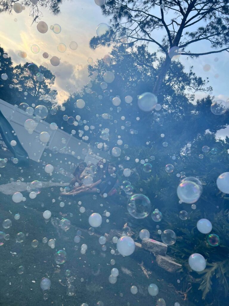 A hazy outdoor scene filled with numerous floating soap bubbles, serving as birthday party add ons, with blurred figures of people visible in the background under trees.