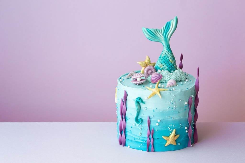 A mermaid slumber party-themed cake with a blue icing base, decorated with seashells, starfish, and a teal mermaid tail on top against a light pink background.