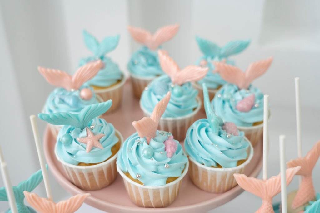 A tray of cupcakes decorated with blue frosting and topped with pink and blue fondant seashells and starfish, perfect for a Mermaid Slumber Party, on a pink background.