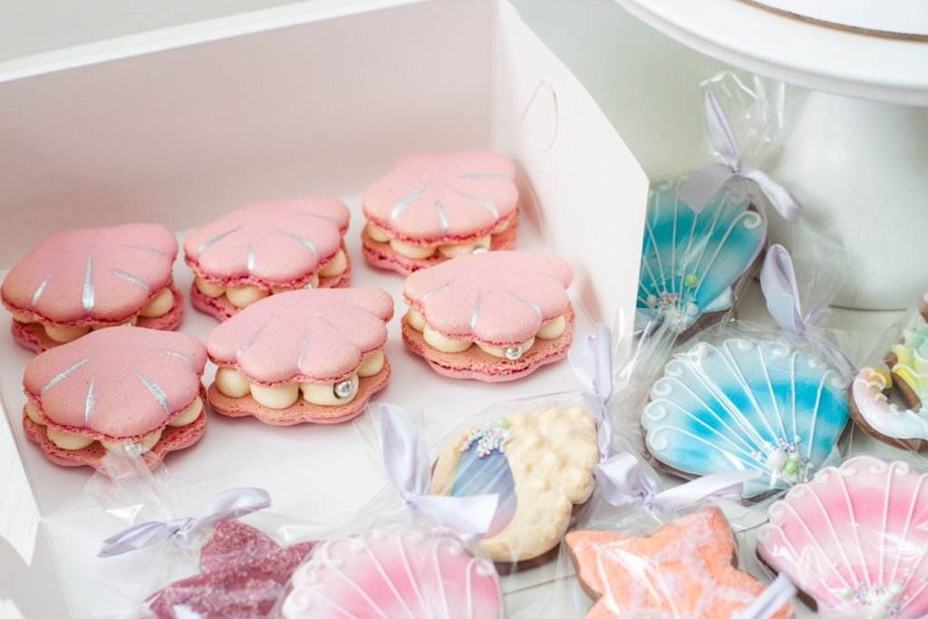 Box of pink seashell-shaped macarons and blue shell-shaped cookies on a table for a mermaid slumber party.