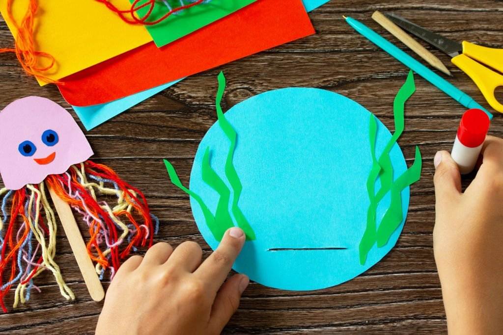 Child's hands crafting with paper, scissors, and glue on a wooden table, making a blue paper circle with green strips for a Summer Slumber Party.