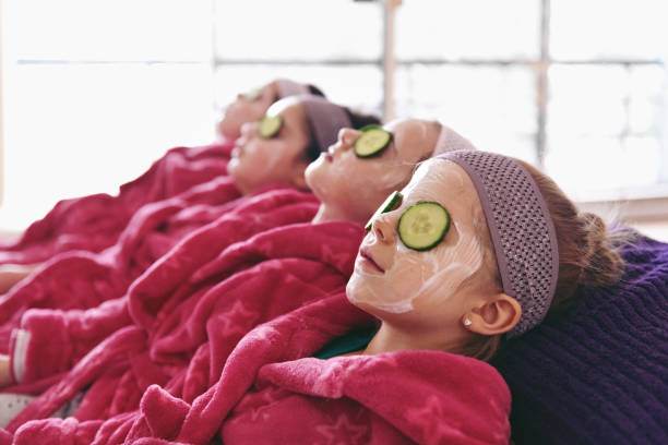 Three people relaxing with facial masks and cucumber slices on their eyes, lying in a row covered with pink blankets as part of birthday party add-ons.