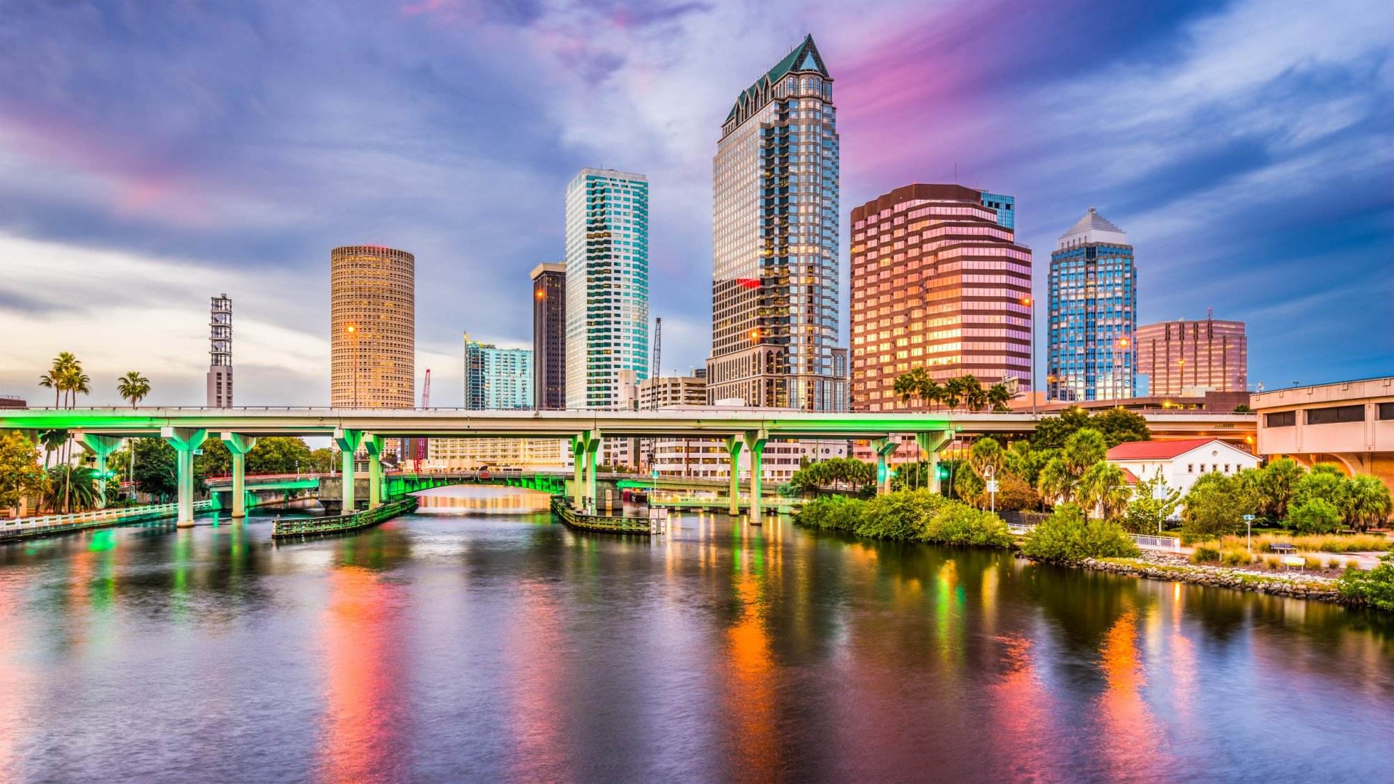 Panoramic view of a vibrant event tampa party rentals and a city skyline with a sunset, featuring modern skyscrapers and a green-lit bridge over a reflective river during a unique slumber party.