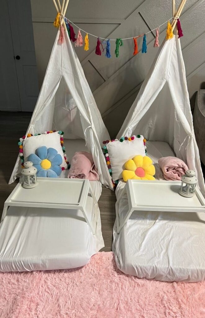 Two children's teepee tents designed for a magical sleepover, with colorful decorations, pillows, and plush blankets inside a room, each tent containing a small lantern on a tray.