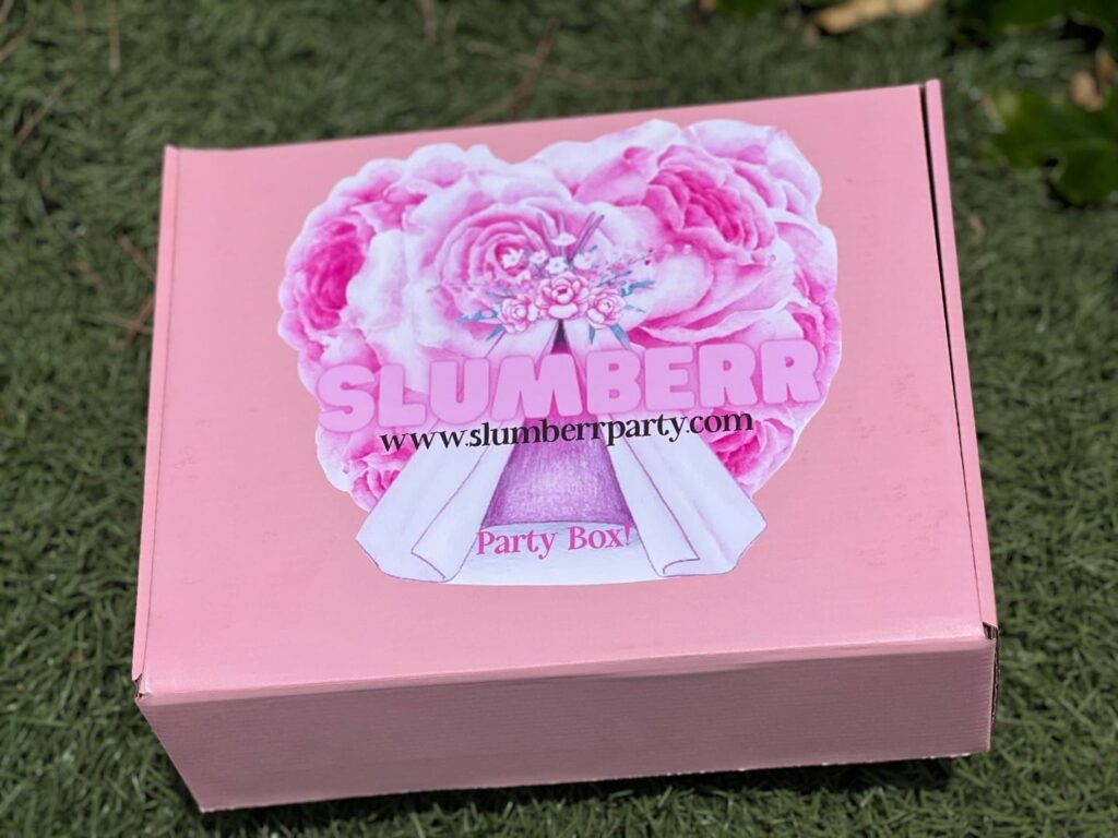 A pink Slumber Party Box with a floral design and the words "Fun Delivered" on top, placed on grass.
