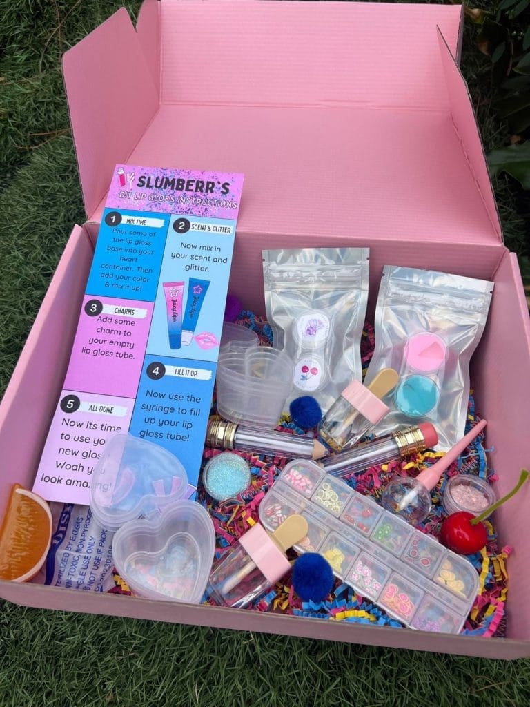Open pink gift box on grass containing various colorful crafting supplies and children's beauty products for an at-home pampering kit, including glitter, beads, and syringe for crafting.