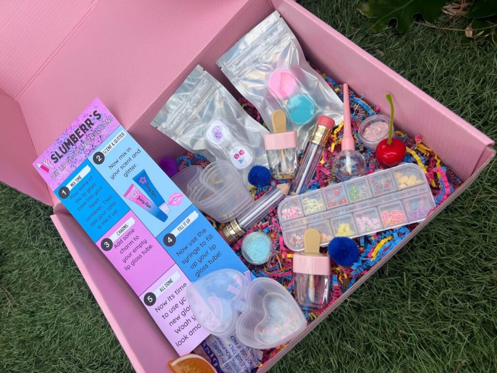 Doorstep Delivery: Open pink gift box on grass filled with colorful craft supplies including glitter, beads, and containers, with informational pamphlets visible.